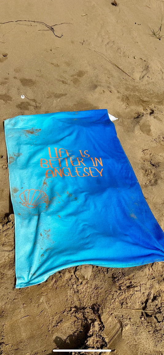 alt=“Vibrant blue microfibre beach towel with the slogan ‘Life is better in Anglesey’ in bold white text – lightweight, quick-drying, and perfect for beach adventures by Shore & Coast”