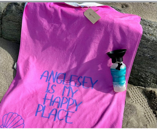 alt=“Pink microfibre beach towel with the slogan ‘Anglesey is my happy place’ in bold white text – lightweight, quick-drying, and perfect for coastal days with Shore & Coast”