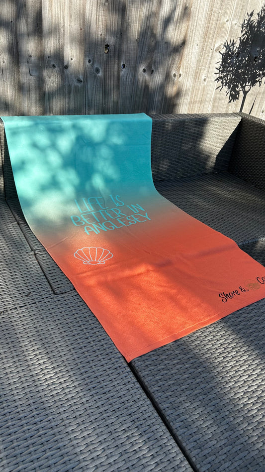 alt=“Mint and orange ombré microfibre beach towel with the slogan ‘Life is better in Anglesey’ in bold white text