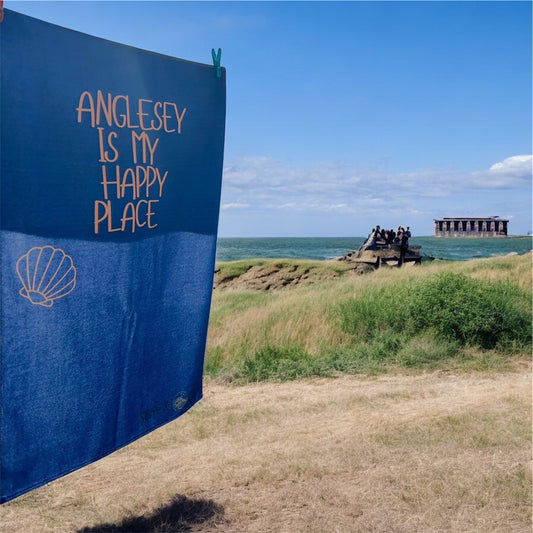 alt=“Navy microfibre beach towel with cotton loops featuring the slogan ‘Anglesey is my happy place’ in bold orange text – quick-drying, absorbent, and perfect for the coast”